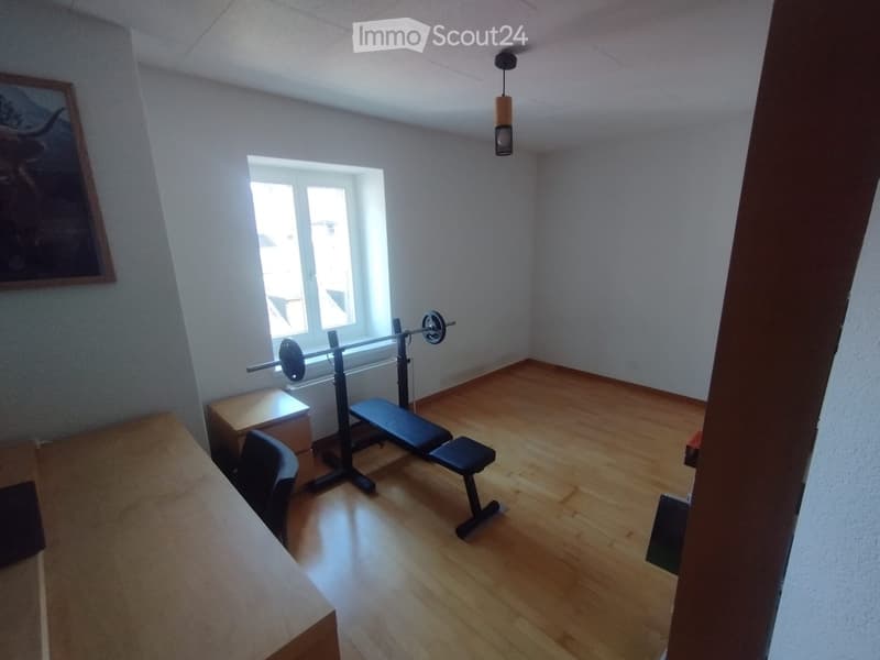 A room with wooden flooring, a barbell with weights, a bench, a stool, a chair, and a small table near the window.