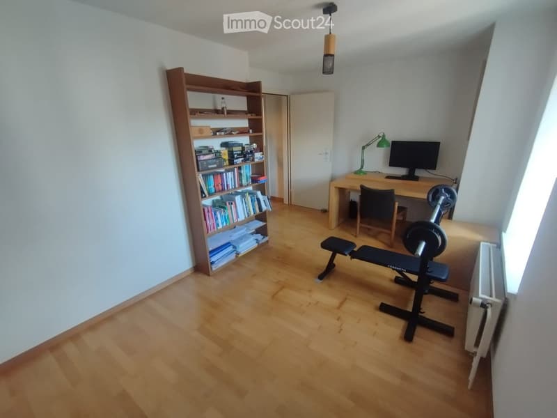 A well-lit room with wooden flooring, a bookshelf filled with books and items, a desk with a computer and chair, and a bench with weights.