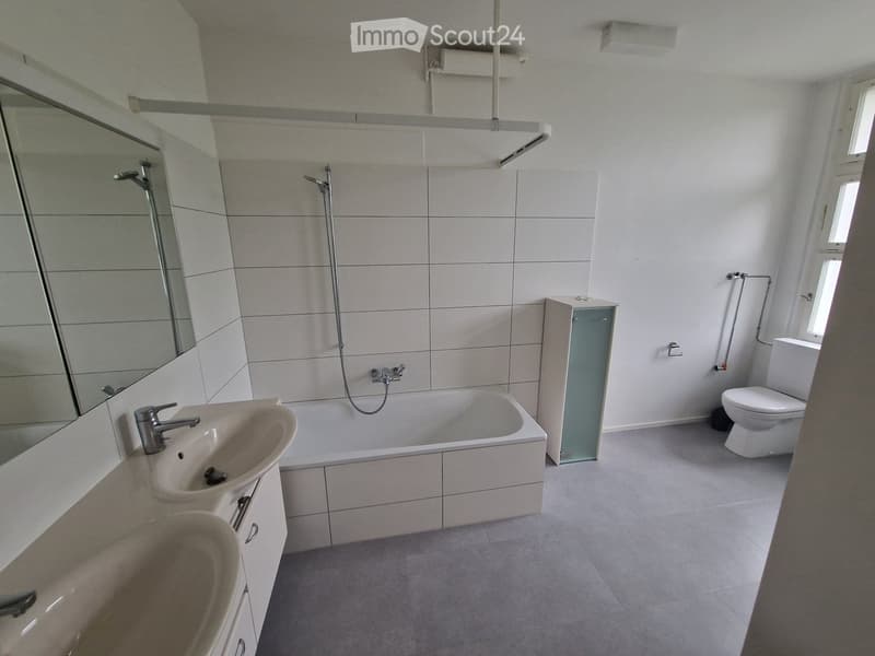 Modern bathroom with white tiles, a double sink, bathtub, toilet, and a large mirror. The space is bright with natural light from a window.
