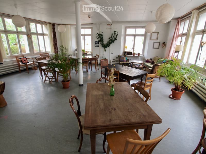 Bright, open room with large windows, wooden tables, chairs, potted plants, and hanging lamps.