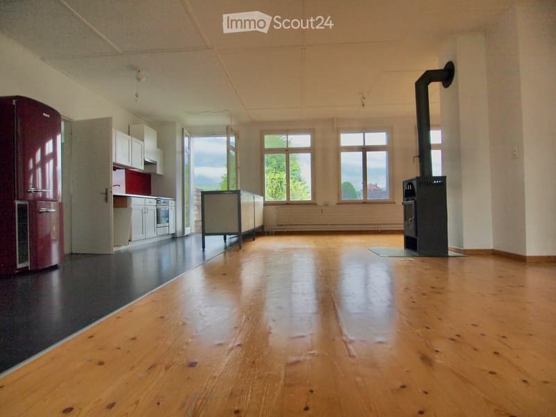 Open-plan room with wooden flooring, a red refrigerator, white cabinets, and a wood stove. Large windows provide natural light.