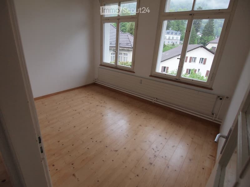 Empty room with wooden flooring, white walls, and large windows offering a view of neighboring houses and greenery.