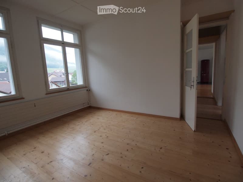 Empty room with wooden flooring, white walls, and large windows offering a view of the outside. The room has a radiator under one window and an open doorway leading to another room.