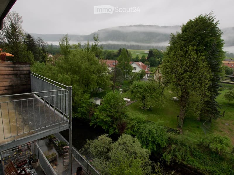 Balcony overlooking a lush green landscape with trees, a small garden area, and distant houses. The view includes a misty valley and hills in the background.