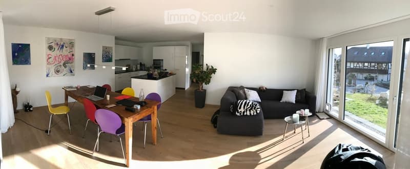 Bright, open-plan living and dining area with wooden flooring, large windows offering a view, a grey sofa with cushions, a coffee table, a dining table with colorful chairs, and a laptop on the table.