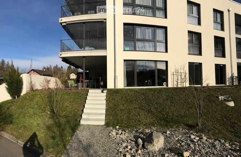 Modern multi-story building with balconies, large windows, raised ground floor, and landscaped garden area.
