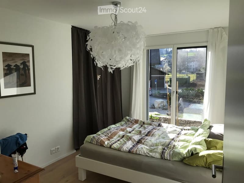 Bright bedroom with a double bed, plaid bedspread, pillows, large window with white curtains, dark brown curtain, and a decorative white chandelier.