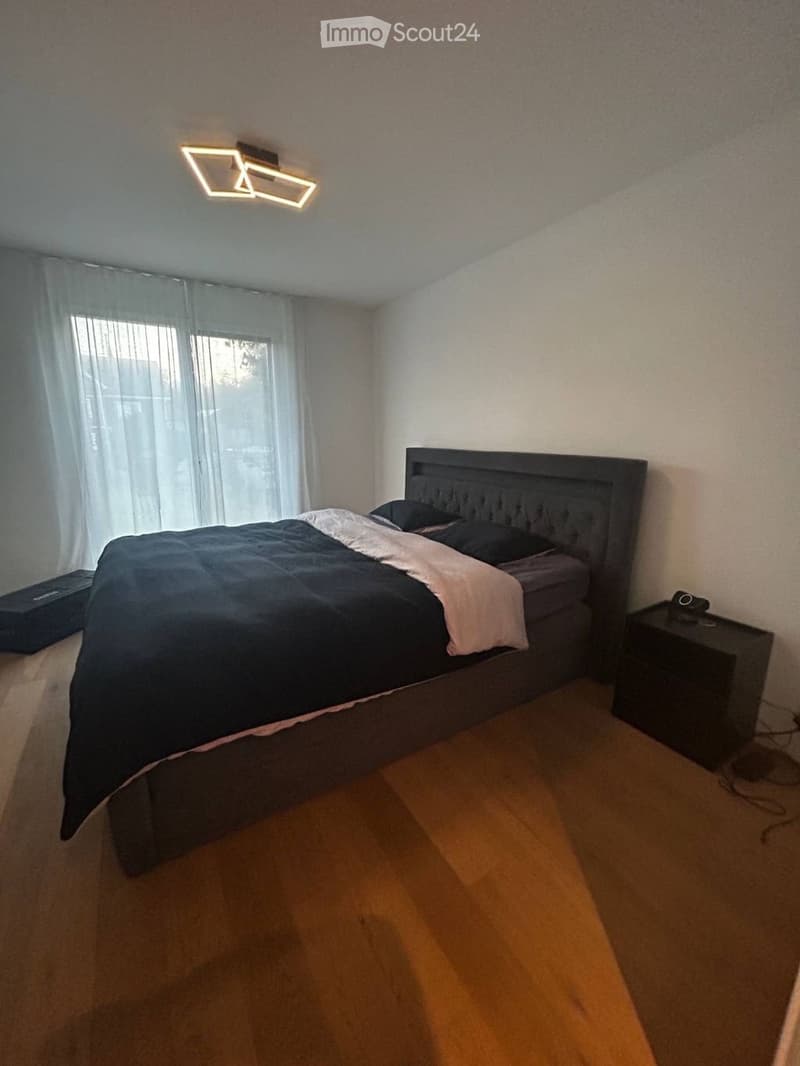 Modern bedroom with wooden flooring, large window with sheer curtains, a dark wooden bed with a tufted headboard, and a matching nightstand.