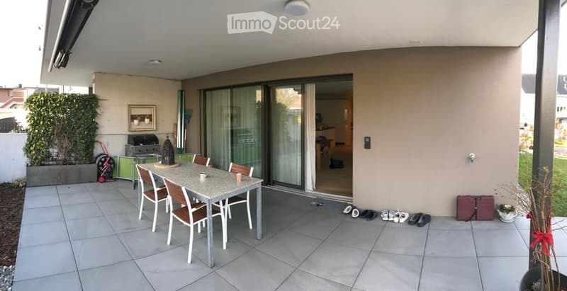 Modern outdoor patio with tiled flooring, a dining table with chairs, a grill, and a small potted plant. The space is connected to the interior via large glass sliding doors.