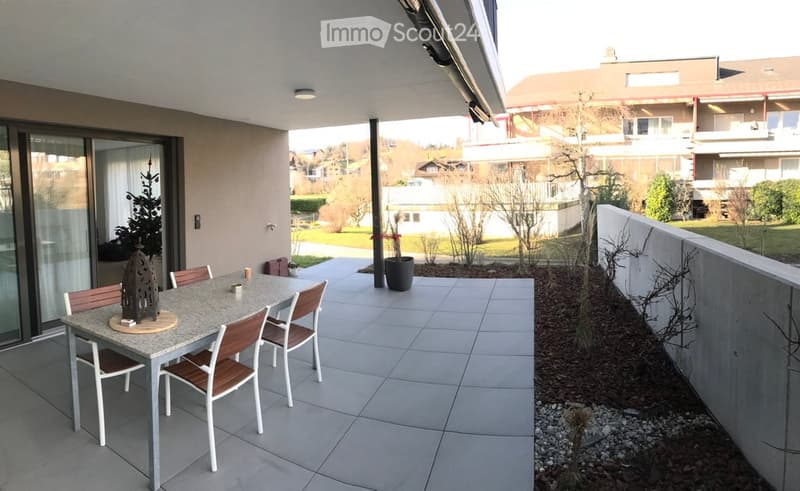 Balcony with a dining table and chairs, small decorative items, and a view of a landscaped garden area with trees and neighboring buildings.