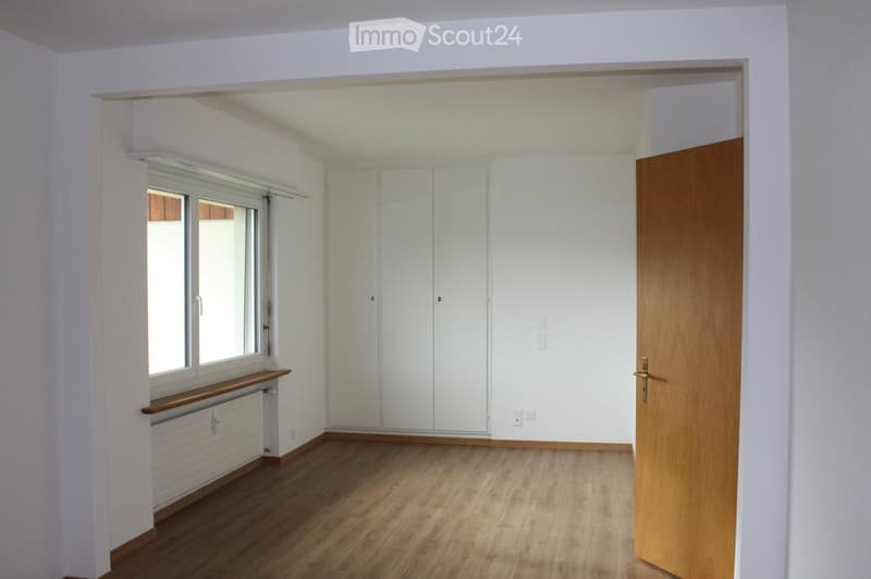 Empty room with wooden flooring, white walls, large window with blinds, built-in white closet, and a wooden door.