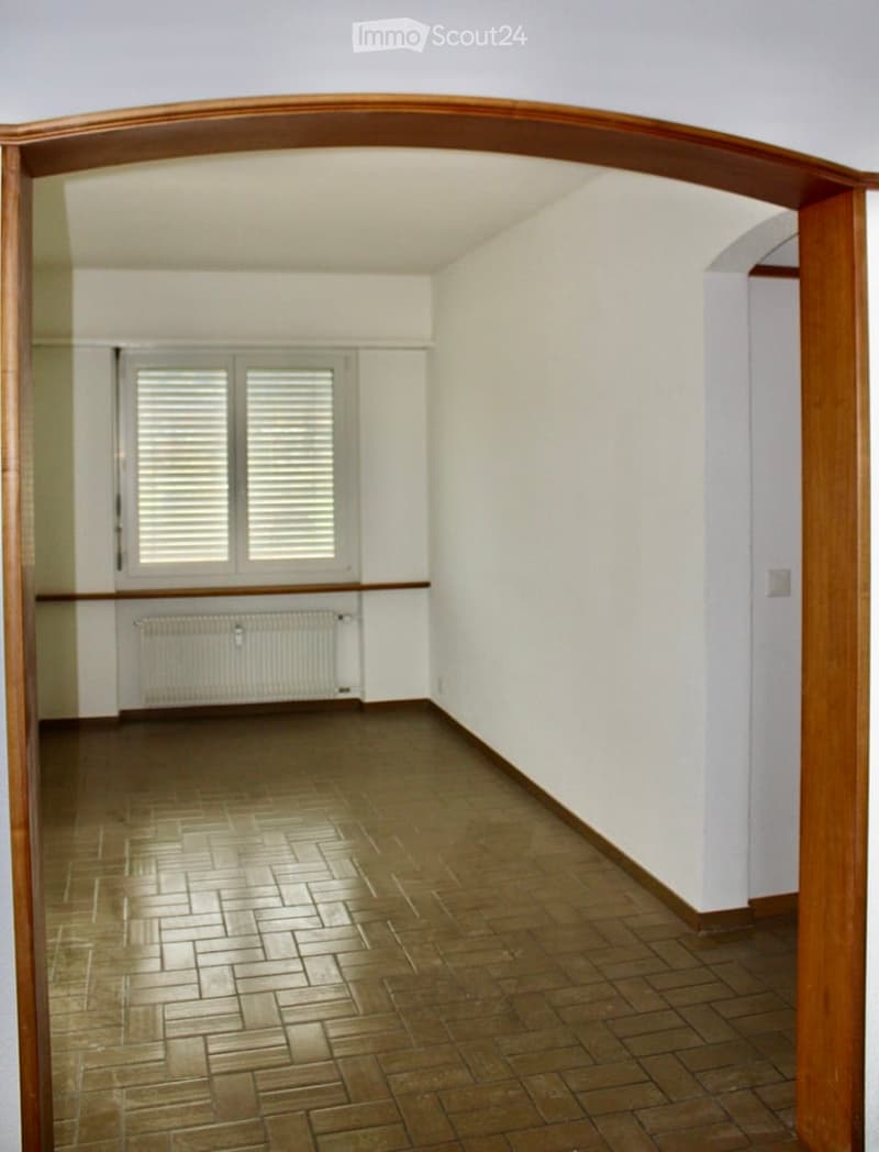Empty room with wooden floor, white walls, large window with shutters, radiator, and wooden archway leading to another space.