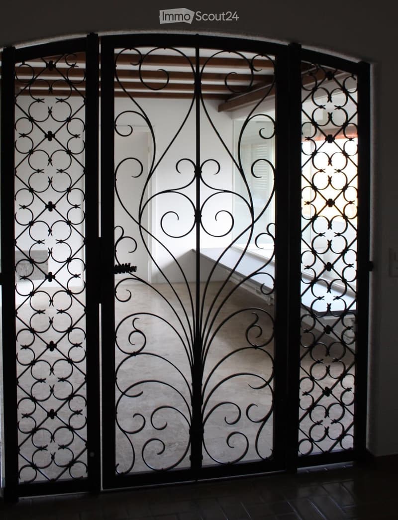Ornate wrought iron gate with intricate scrollwork and floral designs, leading into a well-lit interior space with visible wooden beams and a tiled floor.