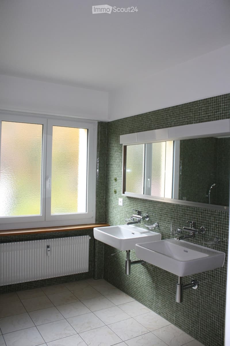 Bathroom with two white sinks, chrome faucets, a large mirror, green tiled walls, and frosted windows.