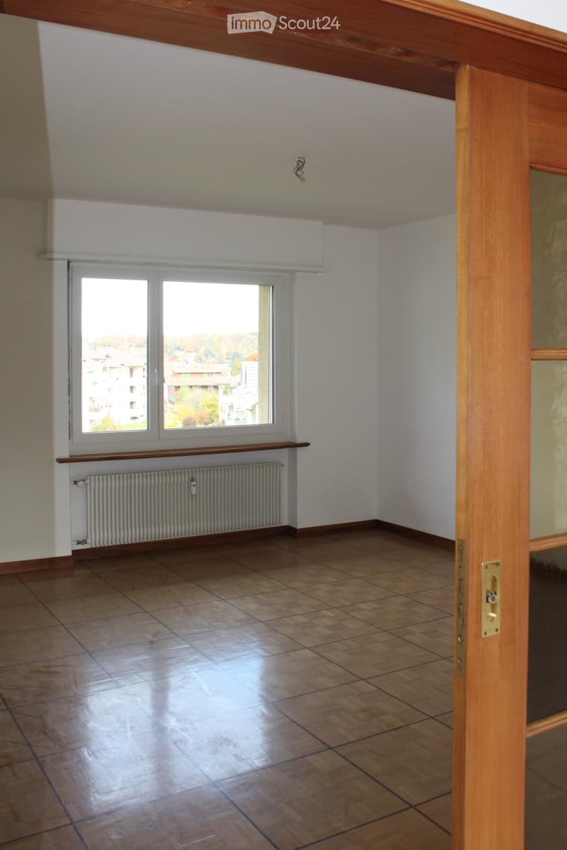 Empty room with wooden flooring, white walls, large window with view, radiator, and wooden door frame.