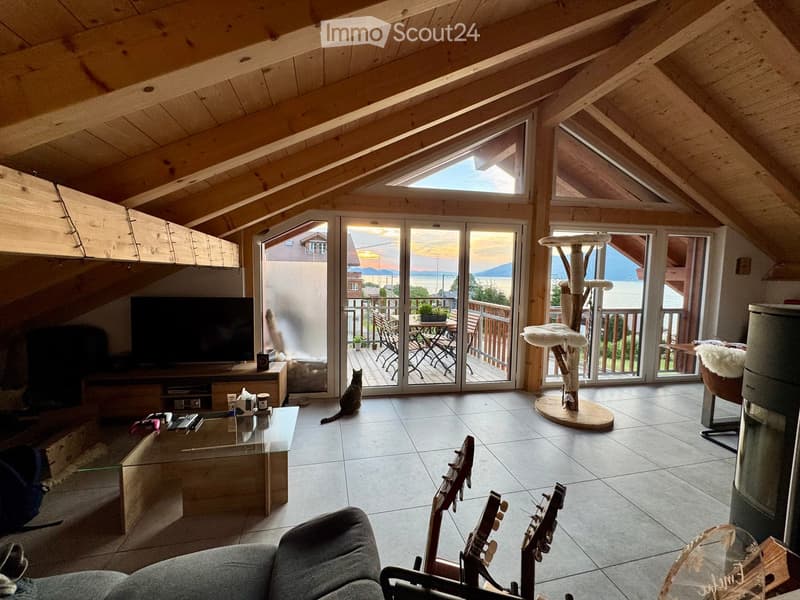 Bright living room with wooden beams, large windows, and a view of the lake. Features a sofa, coffee table, cat tree, guitars, and a television.