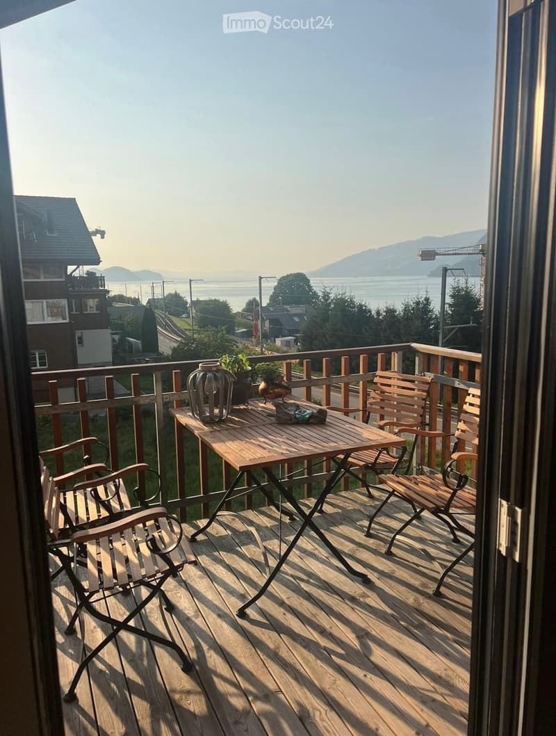 Balcony with wooden decking, table, chairs, vase, and potted plant, overlooking a scenic view of water and distant mountains.