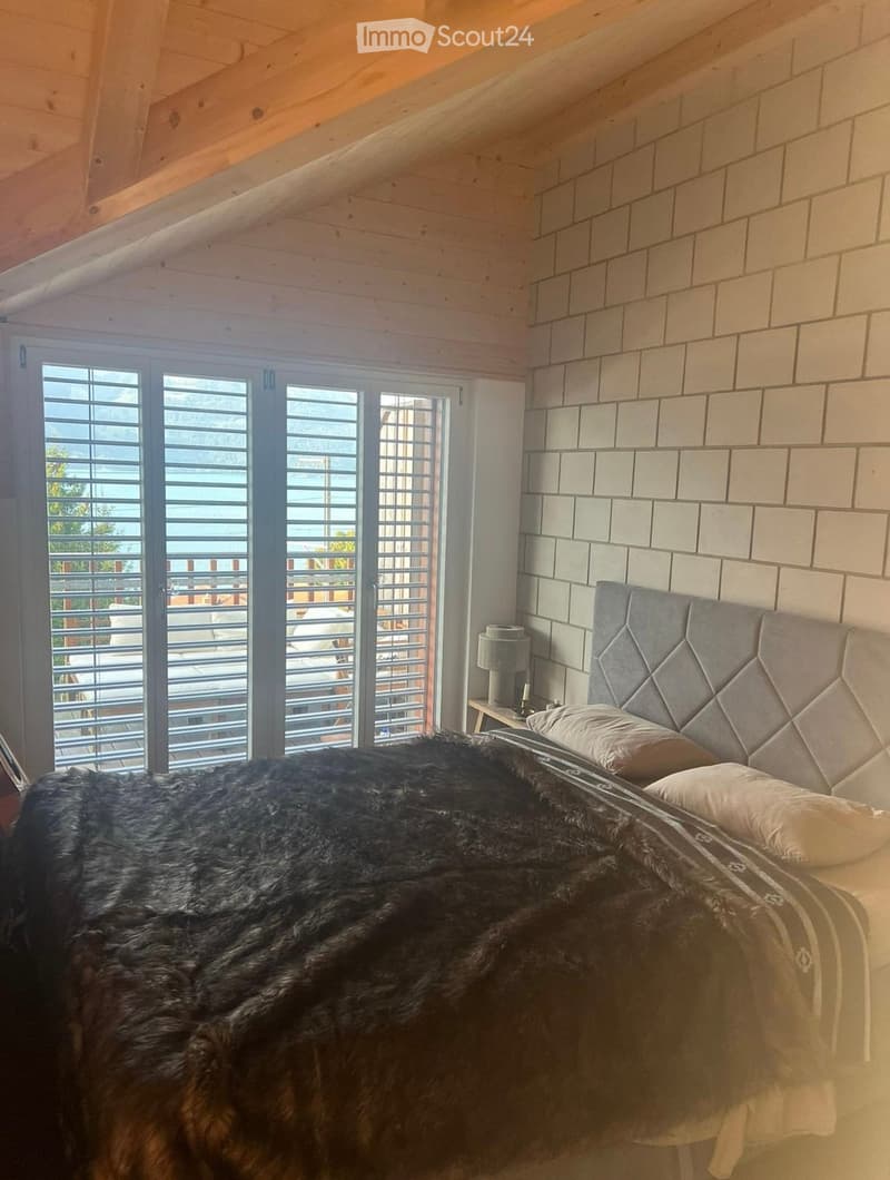 Cozy bedroom with wooden slanted ceiling, large window with shutters, grey upholstered headboard, fur throw, and nightstand with lamp.