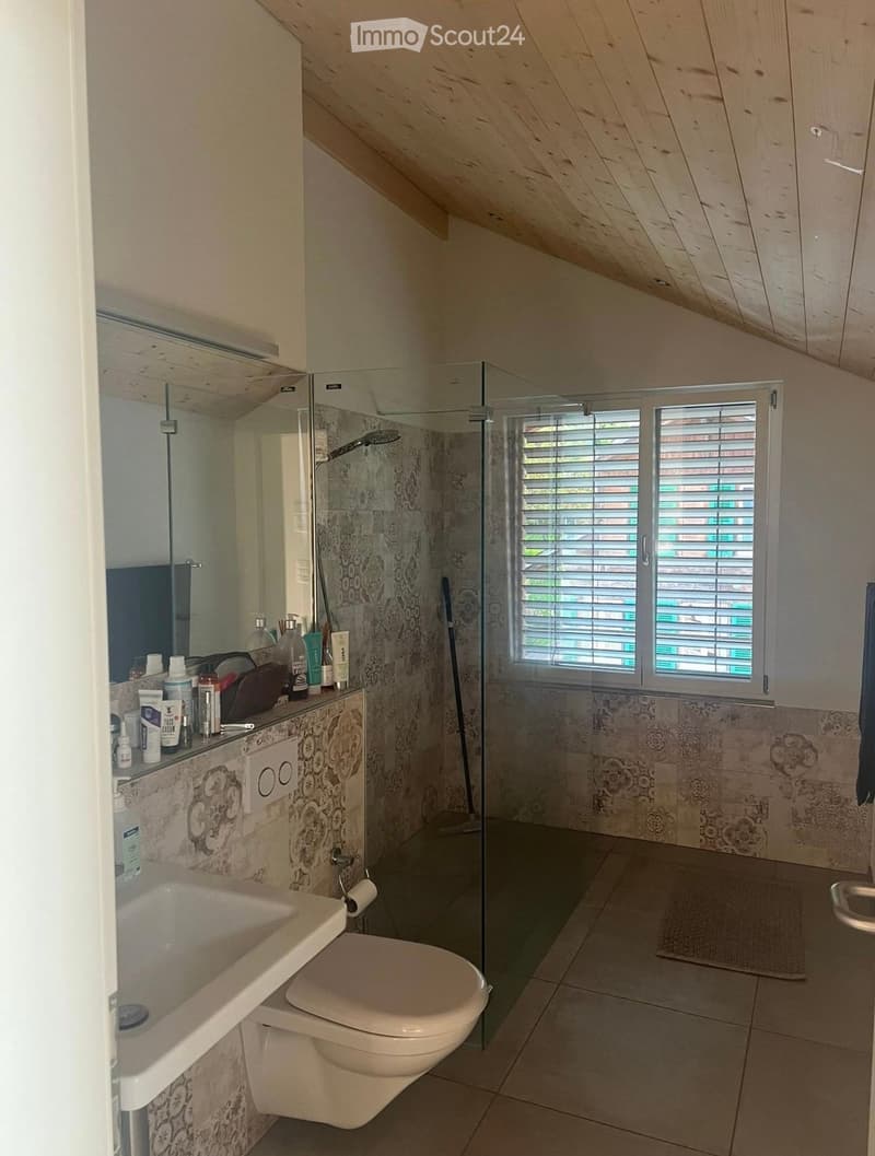 Modern bathroom with a white sink, toilet, and glass-enclosed shower. The walls feature decorative tiles, and the space includes a mirror, various toiletries, and a window with blinds.