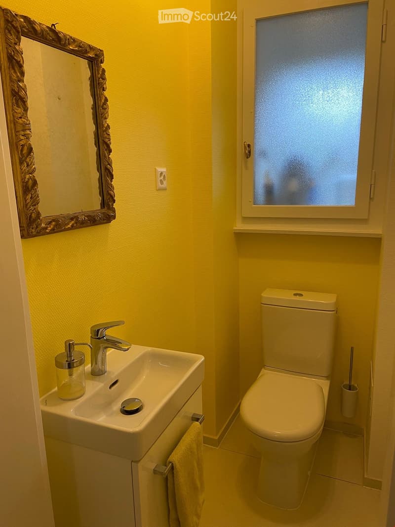 Bright yellow-walled bathroom with a white sink, chrome faucet, and a toilet. A decorative mirror hangs above the sink, and a towel is draped on a rack.