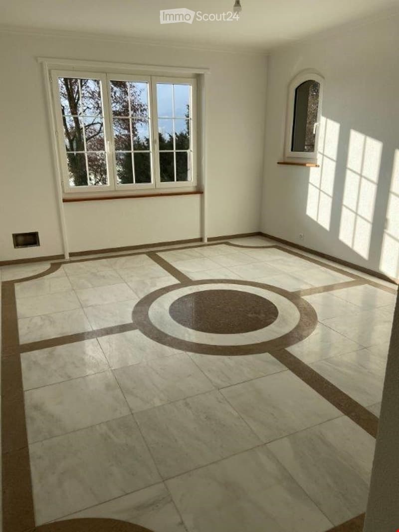 Empty room with large windows, light-colored tiled floor featuring a central circular design, and white walls.