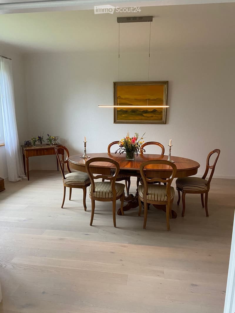 Dining room with a round wooden table surrounded by six chairs, a flower vase in the center, candles on the table, a painting on the wall, and a side table with additional flowers and decorative items.