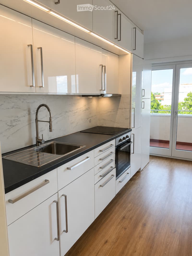 Modern kitchen with white cabinets, black countertops, a stainless steel sink, and a built-in oven. The space features wooden flooring and large windows with a view of greenery.
