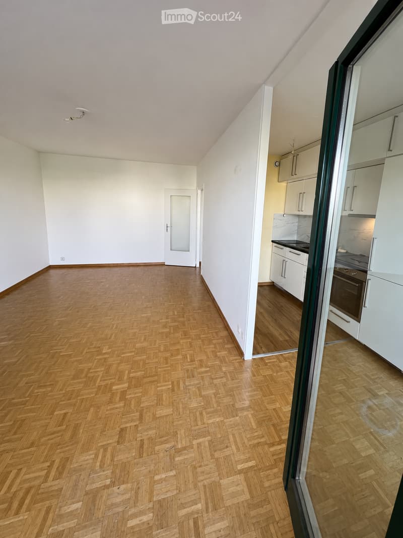 Empty room with wooden herringbone flooring, white walls, and a doorway leading to a kitchen area with white cabinets and appliances.