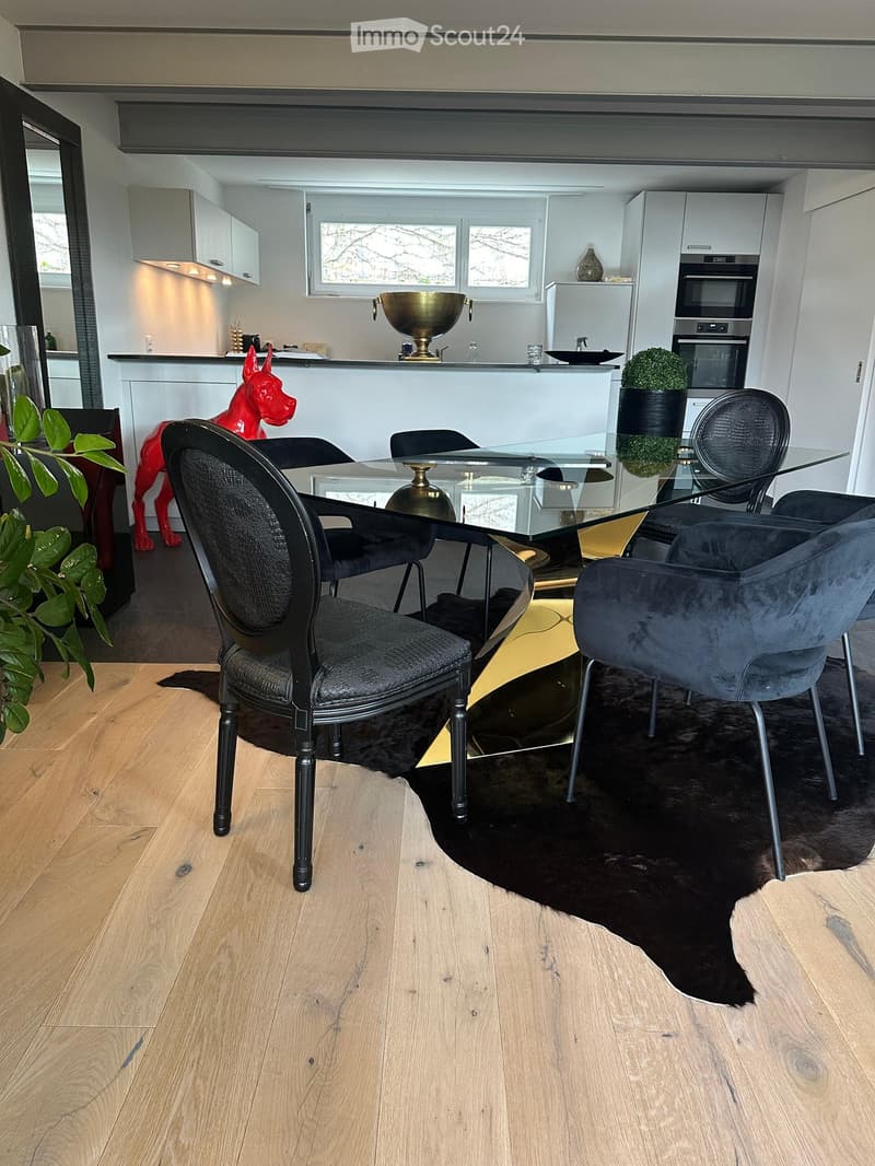 Modern dining area with a glass table, black chairs, and a sleek kitchen in the background, featuring white cabinets, a vase, and a decorative red dog statue.