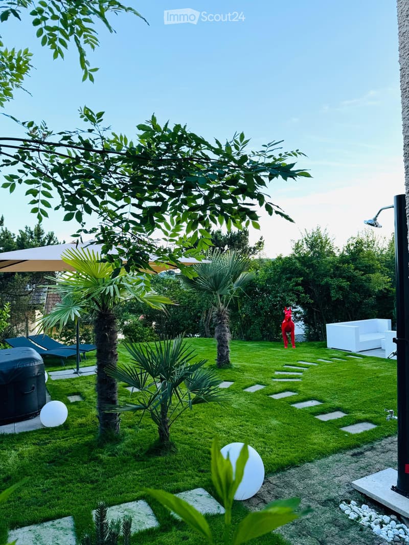 Well-maintained garden with green lawn, palm trees, decorative stones, a white sofa, umbrella, and a hot tub.