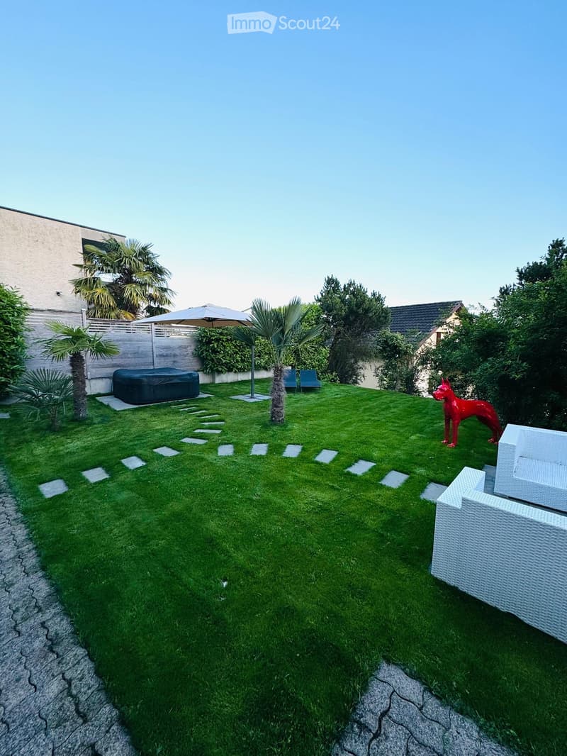 Well-maintained garden with green lawn, white stone stepping stones, a white wicker sofa, a red dog sculpture, a hot tub, and an umbrella near palm trees.
