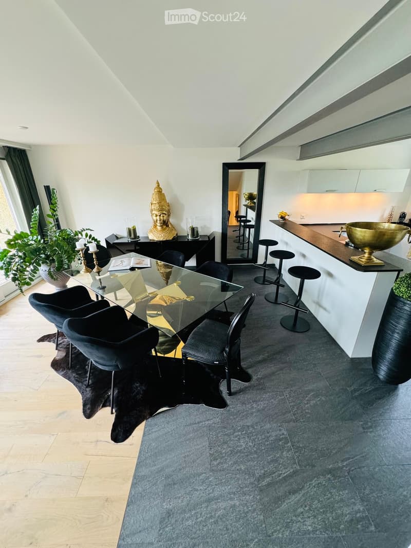 Modern open-plan dining and kitchen area with a glass dining table, black dining chairs, a kitchen counter with stools, a large mirror, and decorative items including a statue and candles.