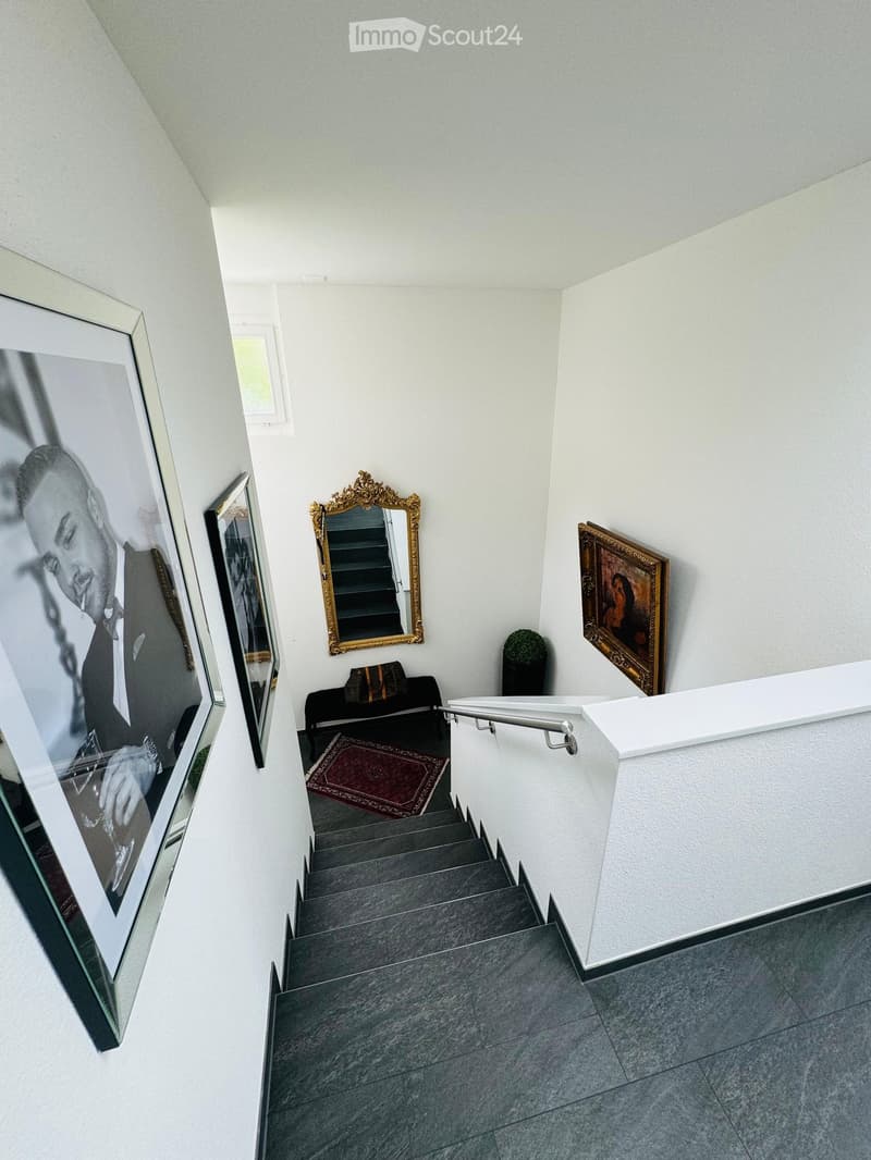 Staircase with grey steps, white walls, framed artwork, small seating area with a chair and table, and a decorative mirror on the wall.
