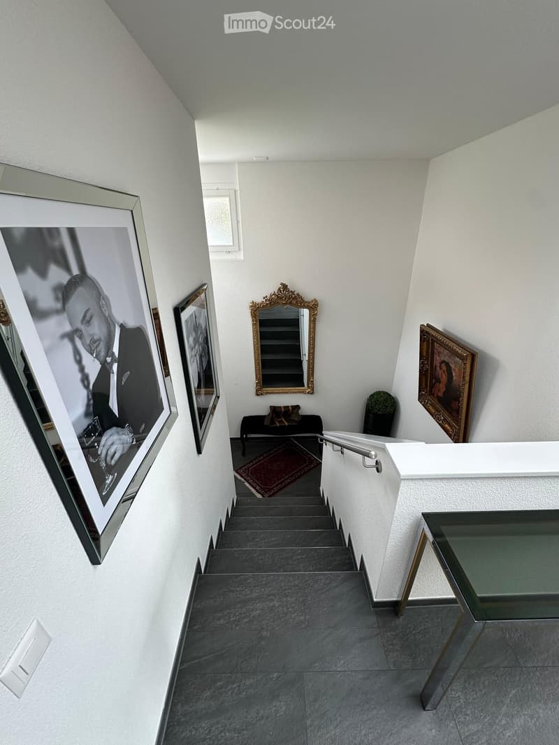 Staircase with grey steps, white walls, framed artwork, small seating area, and a glass table at the bottom.