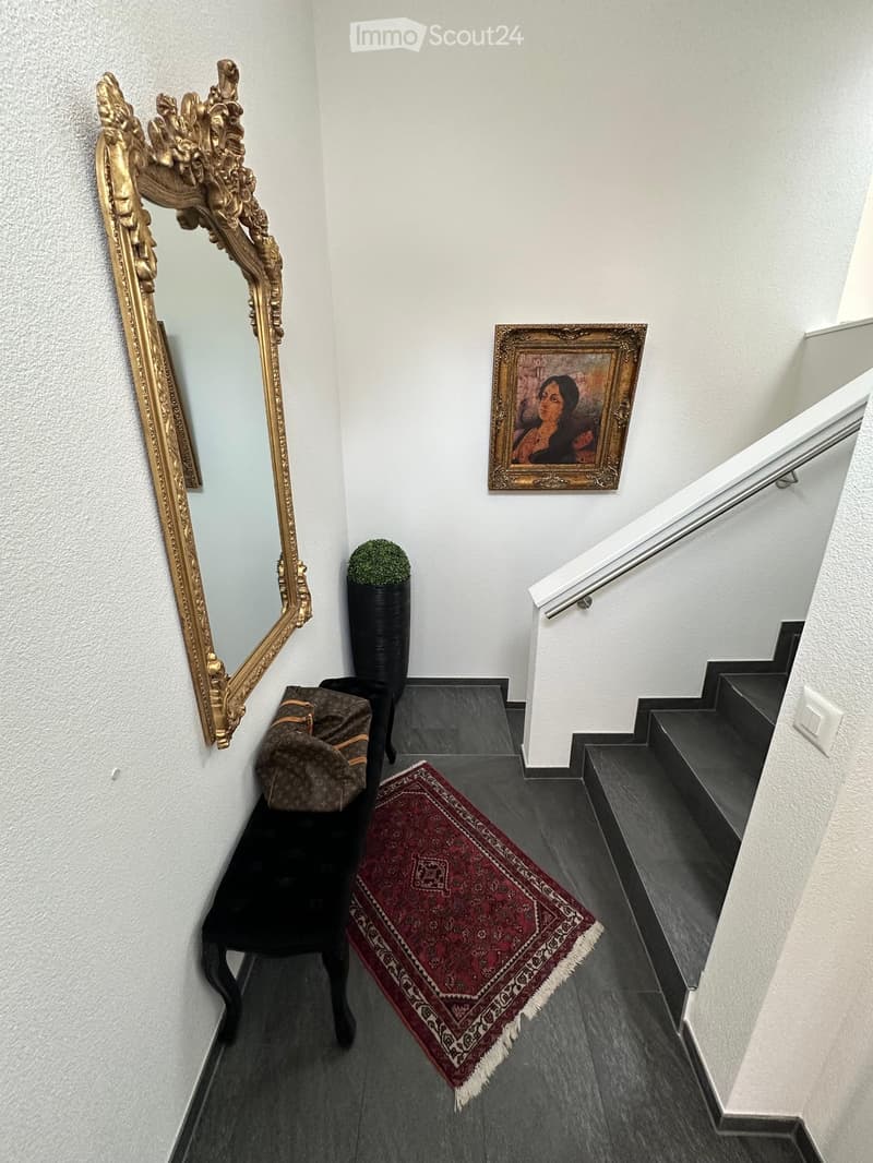 Hallway with dark tiled floor, ornate gold-framed mirror, framed painting, small potted plant, red patterned rug, and a black chair with a bag on it.