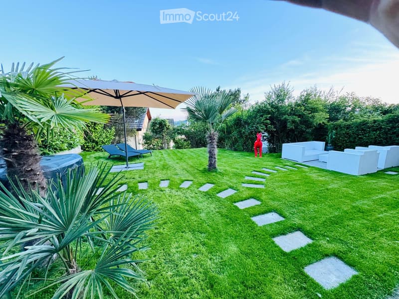 Well-maintained garden with neatly trimmed grass, white outdoor seating, sun loungers, umbrella, decorative pathway, and surrounding greenery.