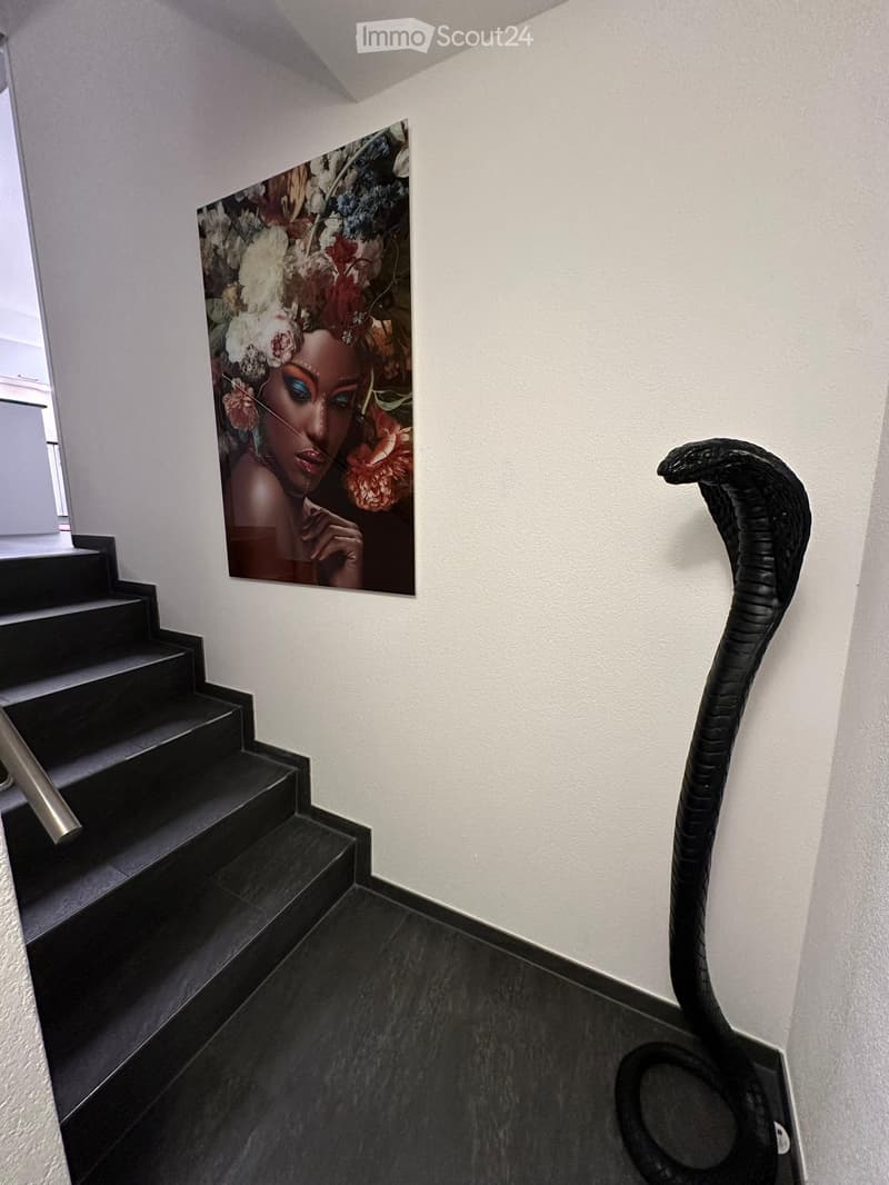 Staircase with black steps and a metal handrail, white walls, a large floral painting, and a black coiled sculpture resembling a snake.