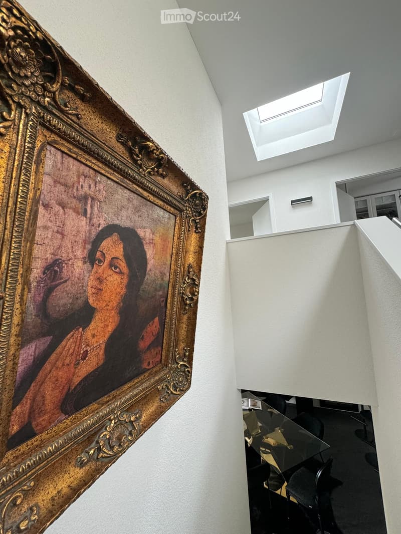 A modern, minimalist room with a large, ornate framed painting on the wall, a glass table, and several black chairs.