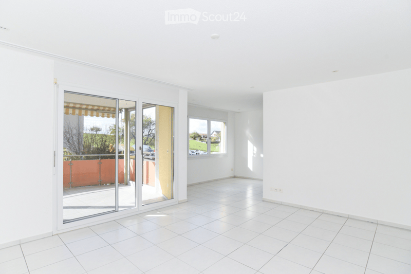 Empty room with white tiled flooring, white walls, large sliding glass doors leading to a balcony, and a window with a view of the outside area.