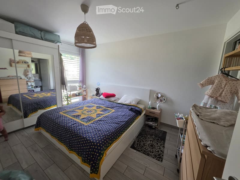 Cozy bedroom with a double bed, nightstand, mirror, wicker pendant light, and a small wardrobe. The room features wooden flooring and a large window with blinds.
