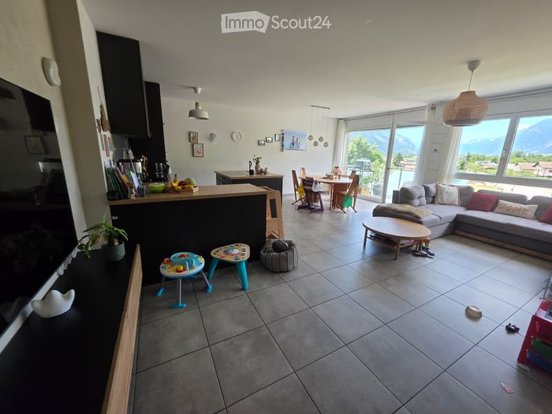 Open-plan living and dining area with a grey sectional sofa, coffee table, dining table with chairs, kitchen island, and a view of mountains through large windows.