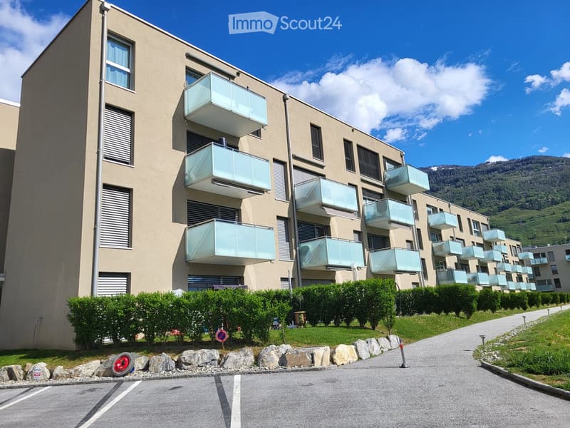 Modern multi-story residential building with glass balconies, surrounded by greenery and a paved driveway.