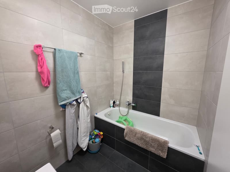 Modern bathroom with a white bathtub, beige tiles, dark grey accents, and towels on a rack. Includes a shower hose, a small trash can, and various toiletries.