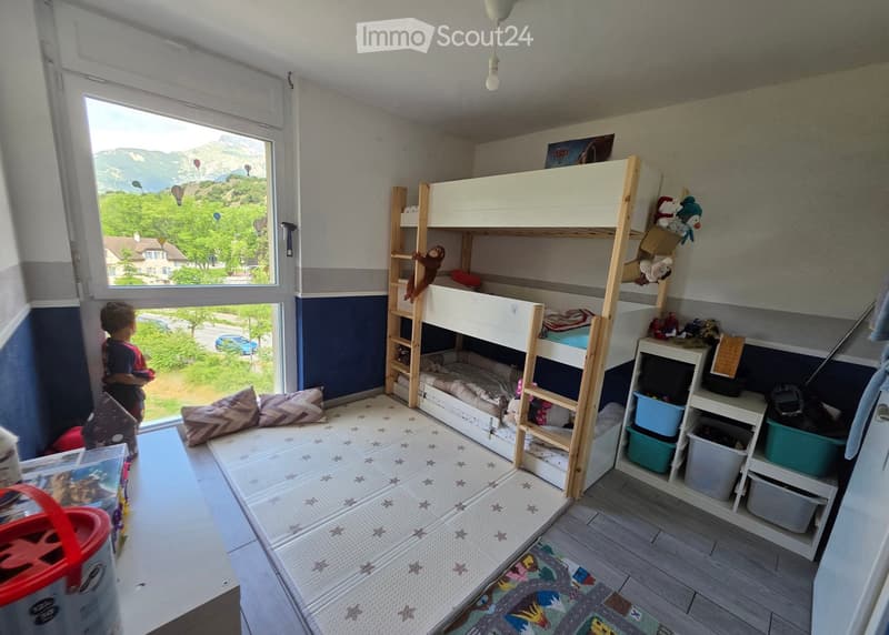 A well-lit bedroom with a bunk bed, shelves, toys, and a view of the mountains through a large window.