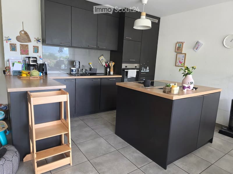 Modern kitchen with dark grey cabinets, wooden countertops, and various appliances including a sink, oven, and coffee maker. There are decorative items such as a vase with flowers, framed pictures, and a clock on the wall.