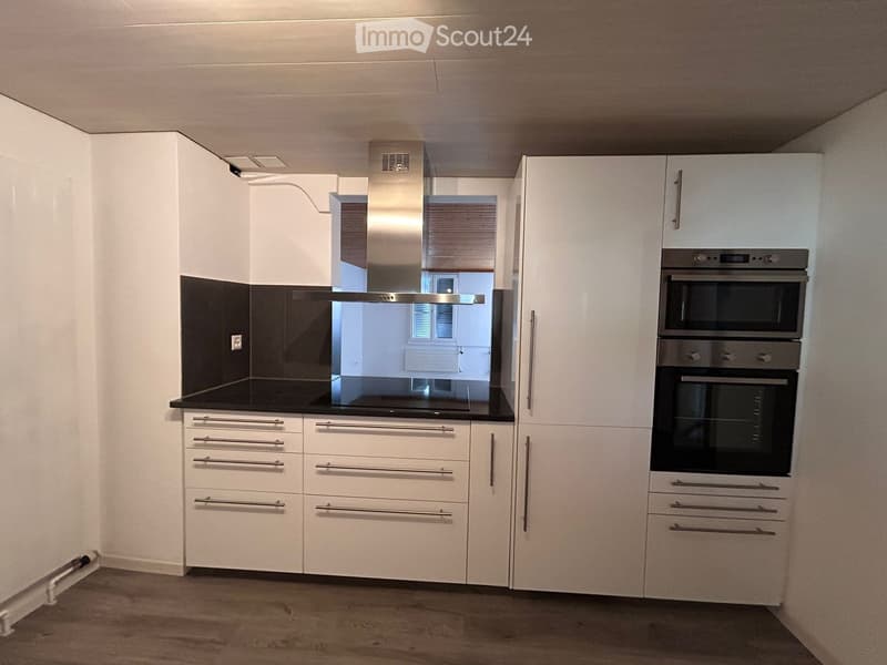 Modern kitchen with white cabinets, stainless steel oven, and range hood. Black countertop and wooden flooring.