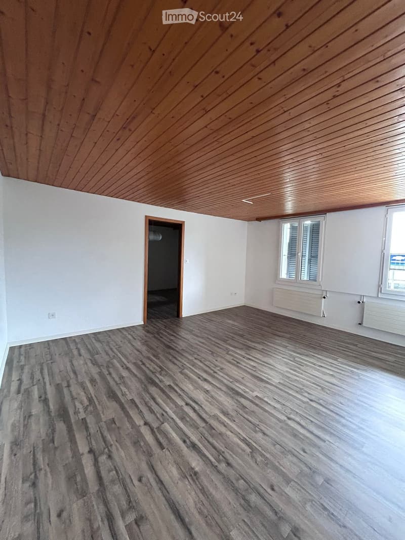 Empty room with wooden flooring, white walls, and a wooden slanted ceiling. Features a window with shutters and a doorway leading to another space.