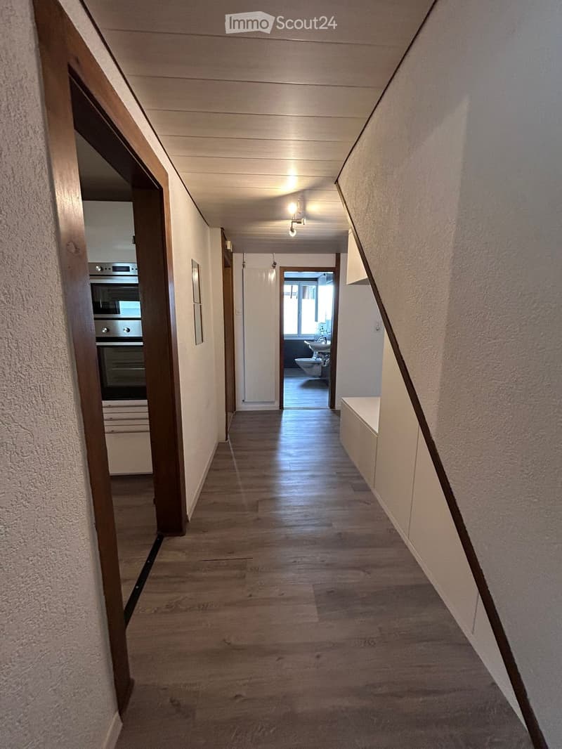A well-lit hallway with wooden flooring, white walls, and recessed ceiling lights. The hallway features an open doorway leading to a bathroom and another doorway to a kitchen area.