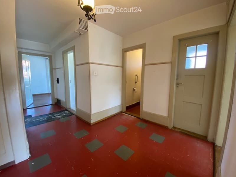 Corridor / Hallway. Empty hallway with red floor, white walls, multiple doors, and a small rug near one entrance.