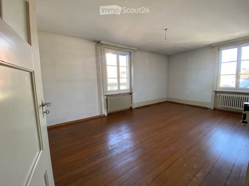 Unfurnished Room. Empty room with wooden flooring, white walls, and two windows with radiators beneath them. The room has a door on the left side and appears to be unfurnished.